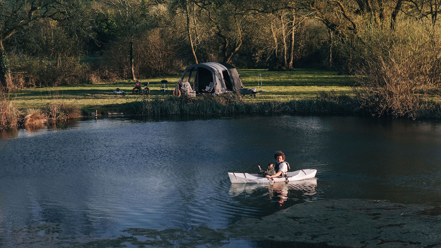 A man in a Kayak with his young child on a camping trip. Their Kampa tent can be seen in the background.